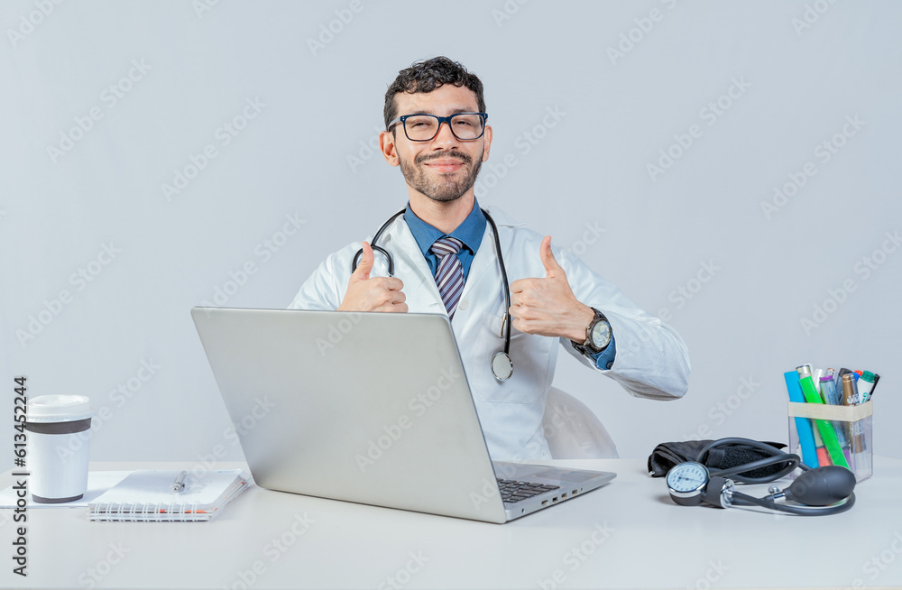 Smiling doctor sitting with laptop giving thumbs up. Male doctor with laptop showing thumbs up in ok gesture. Smiling doctor sitting with laptop giving thumbs up. Male doctor with laptop showing thumbs up in ok gesture.