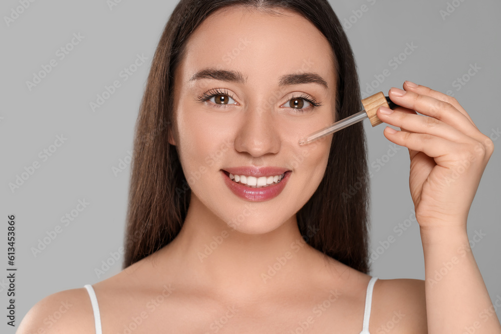 Beautiful young woman applying essential oil onto face on grey background Beautiful young woman applying essential oil onto face on grey background