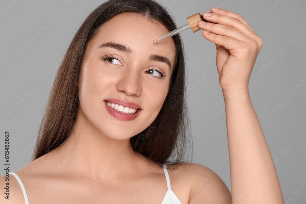Beautiful young woman applying essential oil onto face on grey background Beautiful young woman applying essential oil onto face on grey background