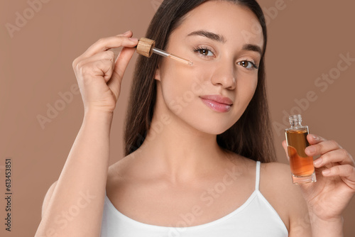 Beautiful woman applying essential oil onto face on brown background