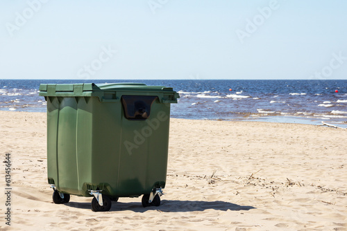  A green trash can is placed on the seashore on the sand