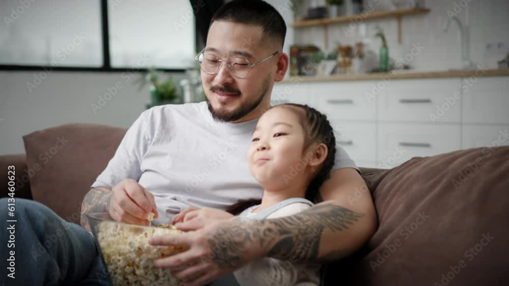 Loving Dad Hugging his Daughter Sitting on Couch at Home. Asian family eating popcorn and watching TV together. Loving Dad Hugging his Daughter Sitting on Couch at Home. Asian family eating popcorn and watching TV together.