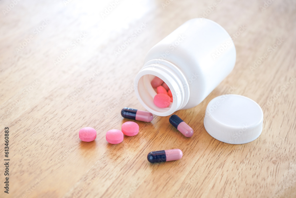 Pink tablets and Capsule spilling from white plastic bottle on wood desk with selective focus. 