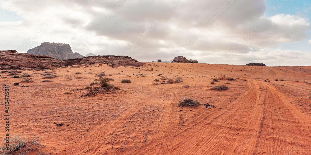 Naklejka premium Rocky massifs on red orange sand desert, vehicle tracks ground, cloudy sky in background, typical scenery in Wadi Rum, Jordan