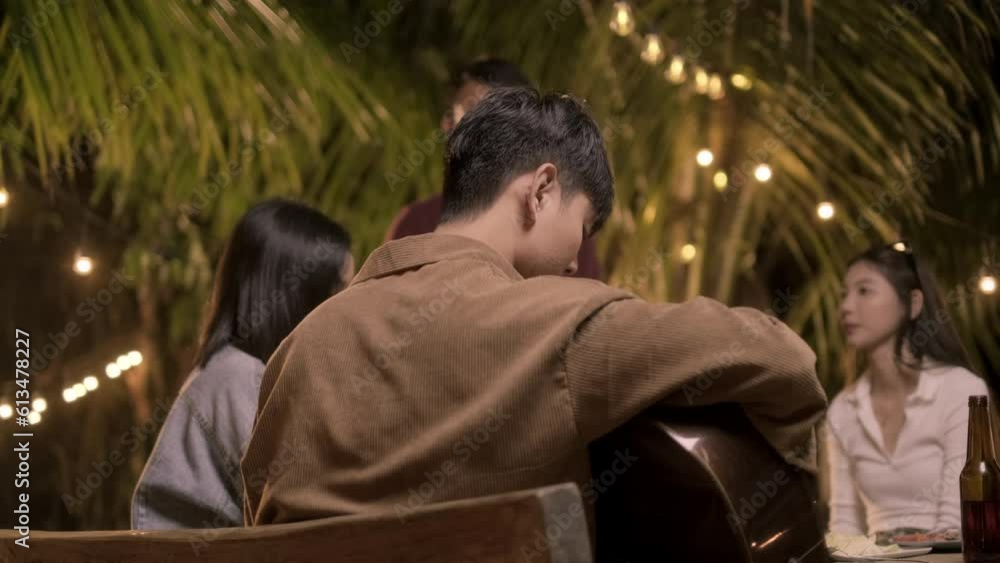 Behind A man playing acoustic guitar and sing song with group friend having fun to music and drinking together outdoor party celebrating. Behind A man playing acoustic guitar and sing song with group friend having fun to music and drinking together outdoor party celebrating.