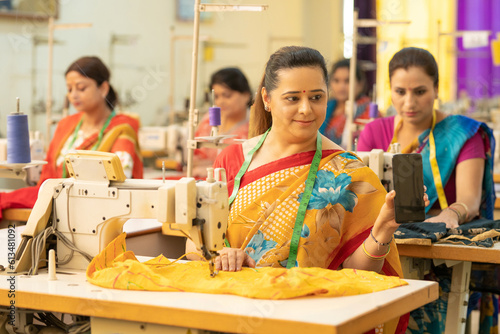 Indian woman showing smartphone screen while working on sewing machine at factory.