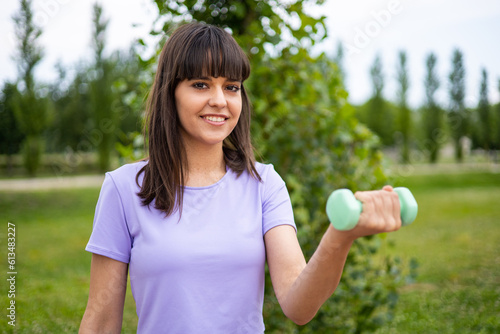 Young woman doing weightlifting sport outdoors. Female fitness exercises with dumbbells in an urban park for strong and healthy muscles. Beautiful lady training weights to get fit. Selective focus.