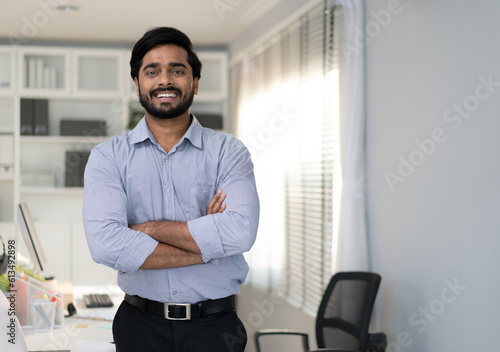Portrait of indian businessman happy standing arms crossed in modern office. Confident young smart bearded man entrepreneur looking at camera smiling. Friendly india ethnic manager work in workplace.