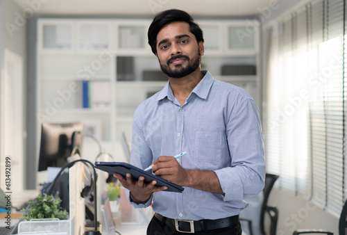 Portrait of indian businessman standing in modern office using tablet computer. Confident smart bearded man entrepreneur looking at camera smiling. Friendly india ethnic manager work in workplace
