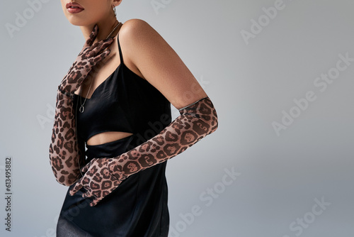 partial view of fashionable woman in black and elegant strap dress, silver necklaces and animal print gloves touching neck on grey background, trendy spring, generation z
