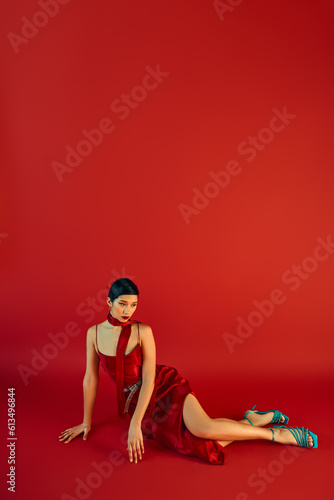 full length of young asian woman with brunette hair and bold makeup, in elegant dress and neckerchief sitting on red background with copy space, spring fashion photography