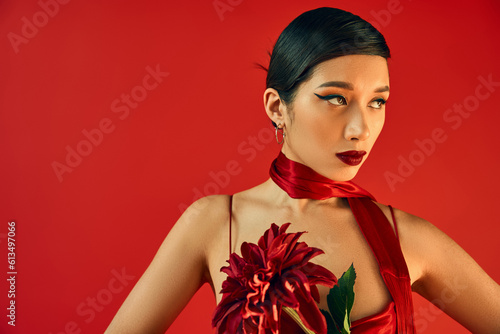 charming asian woman with bold makeup, brunette hair and expressive gaze, in strap dress and neckerchief holding burgundy peopny and looking away on red background, spring fashion photography © LIGHTFIELD STUDIOS
