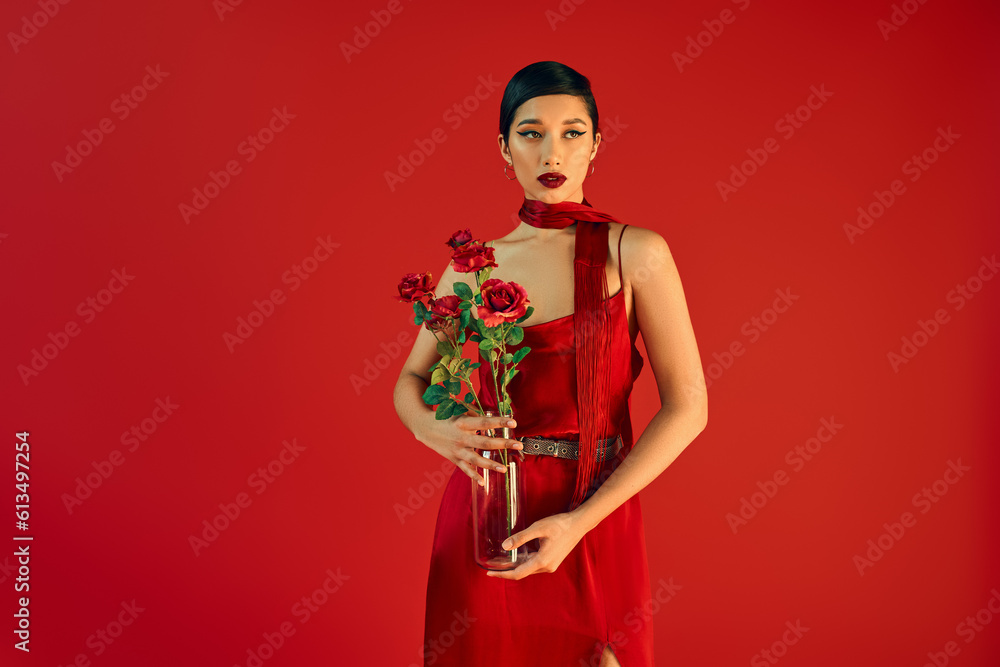 gen z fashion, stylish spring concept, charming asian woman in elegant dress and neckerchief holding glass vase with roses and looking away on red background gen z fashion, stylish spring concept, charming asian woman in elegant dress and neckerchief holding glass vase with roses and looking away on red background