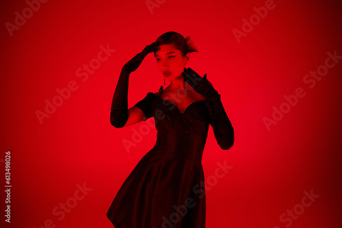 young asian woman in black long gloves and elegant cocktail dress standing in expressive pose and looking away on vibrant background with red lighting effect, youth culture, fashionable spring