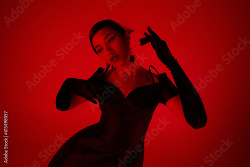 expressive asian woman with brunette hair, in black long gloves and stylish cocktail dress posing on vibrant background with red lighting effect, trendy spring, youth culture concept