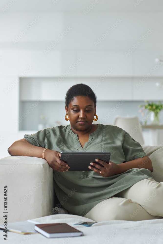 Vertical image of African American woman working online on tablet pc while sitting on sofa in the room at home Vertical image of African American woman working online on tablet pc while sitting on sofa in the room at home