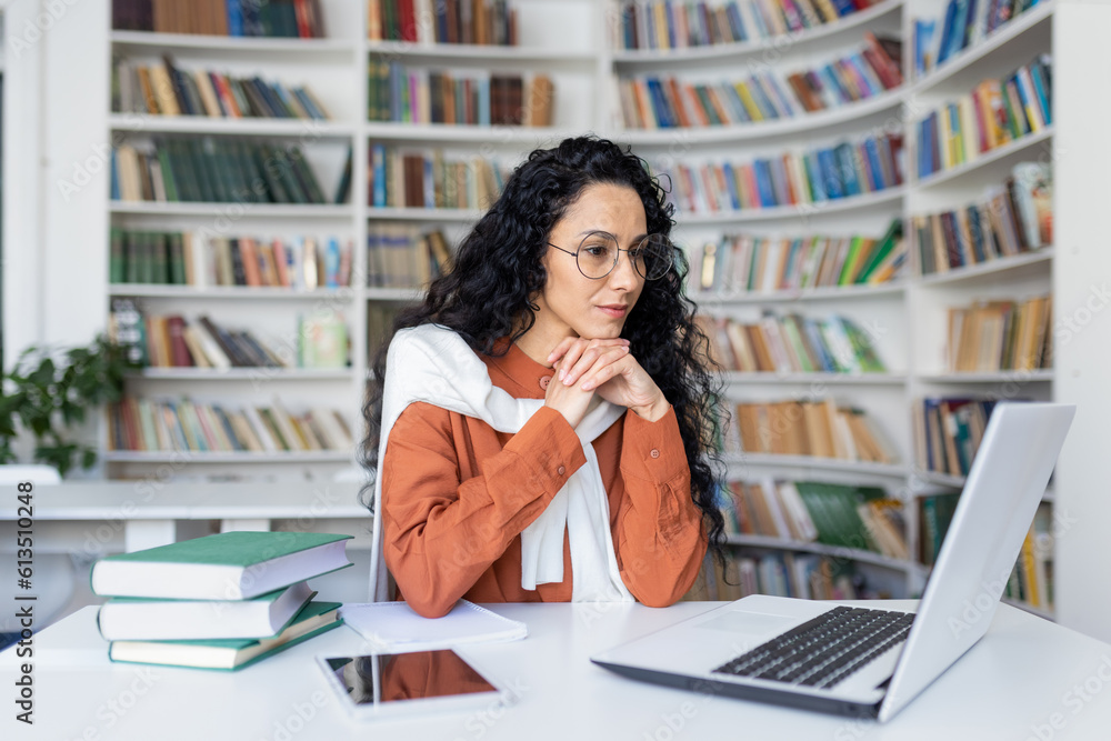 Young serious female student thinking about a solution to an assignment, studying remotely while sitting in a university campus library, Hispanic woman using a laptop to prepare for exams. Young serious female student thinking about a solution to an assignment, studying remotely while sitting in a university campus library, Hispanic woman using a laptop to prepare for exams.