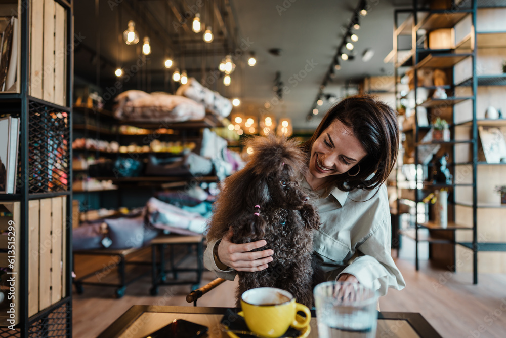 Beautiful and happy young woman sitting in modern pet shop cafe bar and enjoying in fresh coffee together with her adorable brown toy poodle.