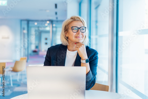 50's confident mature businesswoman dreamy looking at window, middle-aged experienced senior female professional working on laptop in open space office. Female entrepreneur working remotely