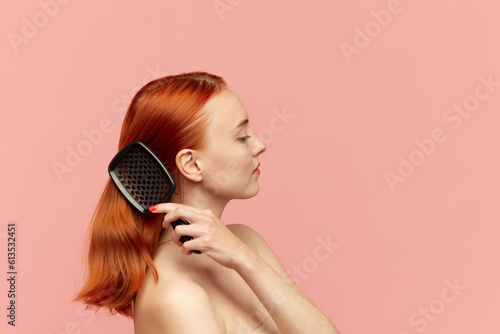 Photography Portrait of charming, redheaded girl with silk, shine hair combing her hair and looking away over pink background