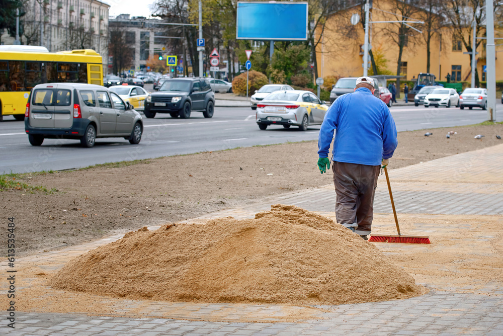 Pile of sand lies on new sidewalk, man seal seams between paving slabs