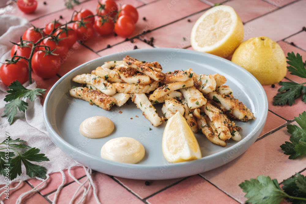 fried squid, fish pieces with white sauce, lemon and garlic. on a pink tiles table, fresh ingredients scattered around