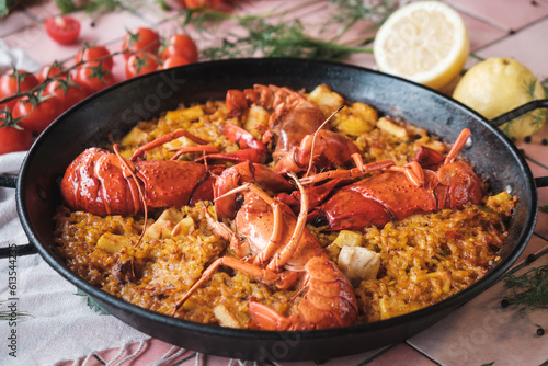 spanish seafood paella with lobster and squid, traditional dish with rice in a hot pot, surrounded by fresh ingredients on a pink background table, close up