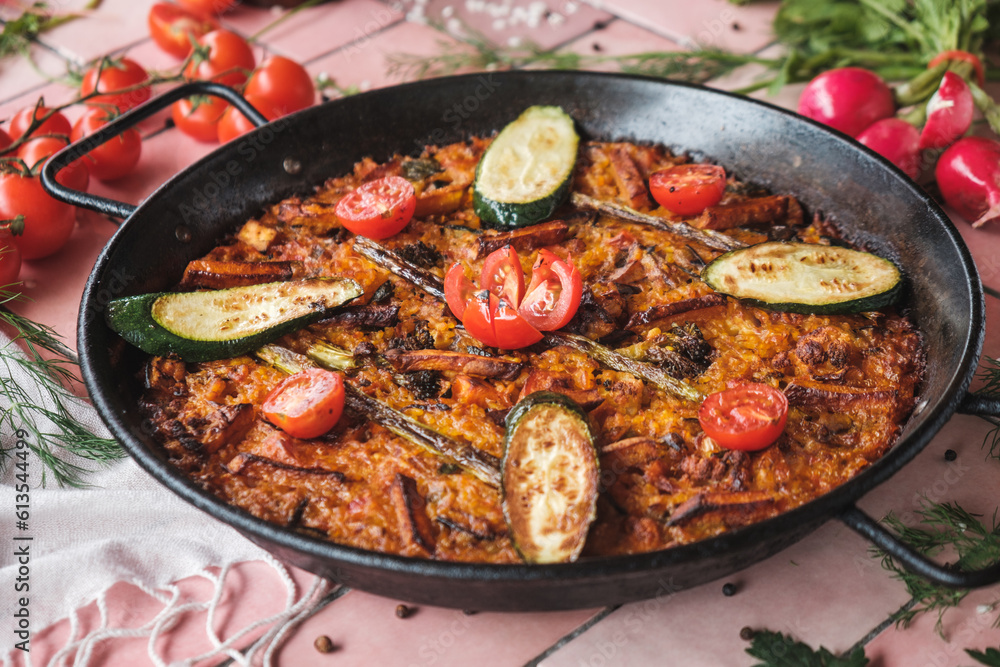 spanish paella with vegetables, zucchini, tomatoes, asparagus, traditional dish with rice in a hot pot, surrounded by fresh ingredients on a pink background table