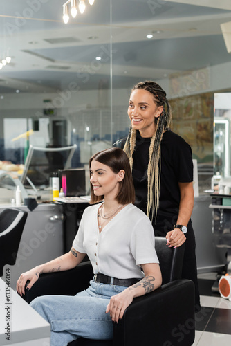 happy hairdresser and female client in beauty salon, cheerful beauty worker with braids standing near tattooed woman, discussing hair treatment, hair extension, customer satisfaction