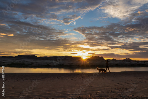 Silhouette of woman walking with her dog along the shoreline of Lake Powell on Lone Rock Beach in Wahweap, Glen Canyon Recreation Area, Page, Utah, USA. Sand beach on wild campground. Red orange sky