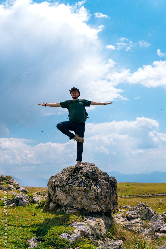 A male traveler in a hat and backpack stands on a rock boulder in yoga pose. Svaneti region, Georgia. Summer day in the mountains of the Caucasus. Tourism and travel. Vertical photo A male traveler in a hat and backpack stands on a rock boulder in yoga pose. Svaneti region, Georgia. Summer day in the mountains of the Caucasus. Tourism and travel. Vertical photo
