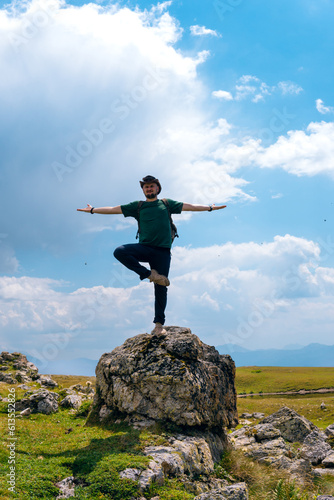 A male traveler in a hat and backpack stands on a rock boulder in yoga pose. Svaneti region, Georgia. Summer day in the mountains of the Caucasus. Tourism and travel. Vertical photo
