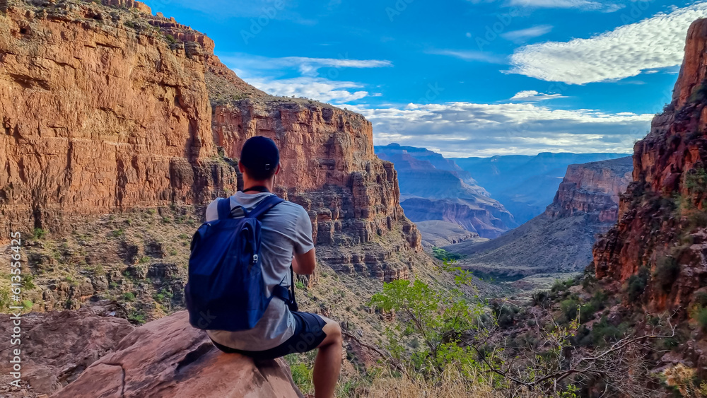 Fototapeta premium Rear view of man with backpack sitting on rock along Bright Angel trail with aerial overlook of South Rim of Grand Canyon National Park, Arizona, USA, America. Amazing vista after sunrise in summer