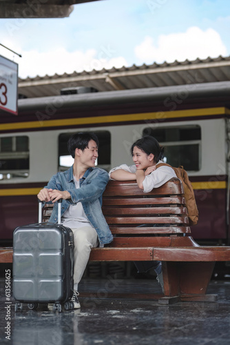 A lovely young Asian tourist couple with their suitcases enjoys talking on a bench on the platform before boarding the train together. Honeymoon trip, Summer vacation, Holidays