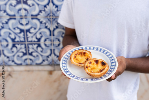 Plate with Pastel de Nata, Portugal\'s traditional sweet dessert, egg custard tart pastry, in hands of young African man in front of a wall with azulejo tiles in Lisbon
