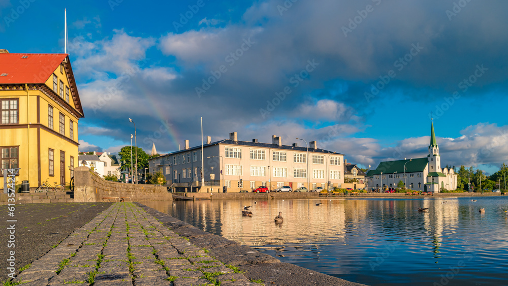 Obraz na płótnie Historical and touristic downtown in Reykjavik at sunset and rainbow in Iceland. Cityscape at golden hour and blue sky at inner lake around Tjornin city park in the downtown. w salonie