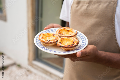 Fototapeta Naklejka Na Ścianę i Meble -  Young African man waiter serving 
 a plate with Portugal's traditional sweet dessert Pastel de Nata, egg custard tart pastry, in the street cafe