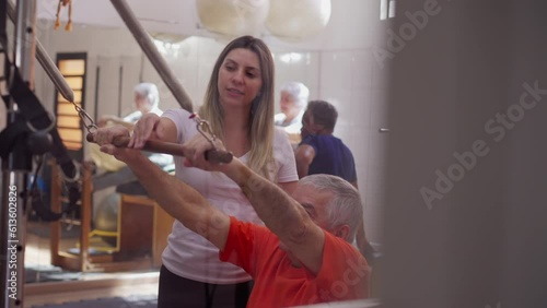 Elderly man exercising in Pilates Studio with the help of a female sport coach. Person using Physiotherapy machine to strengthen muscle and gain flexibility