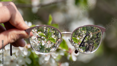 Myopia (shortsightedness) glasses in hand, looking on blooming spring trees garden in focus with blurry background. Nearsighted refractive lenses outdoors in nature. 