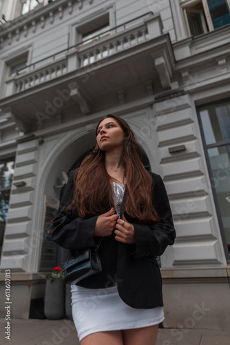 Beautiful serious young fashionable girl in stylish casual clothes with white dress and black blazer and handbag walks in the city near a white vintage building