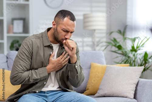 Photographie Young African American man sitting on the couch at home and coughing