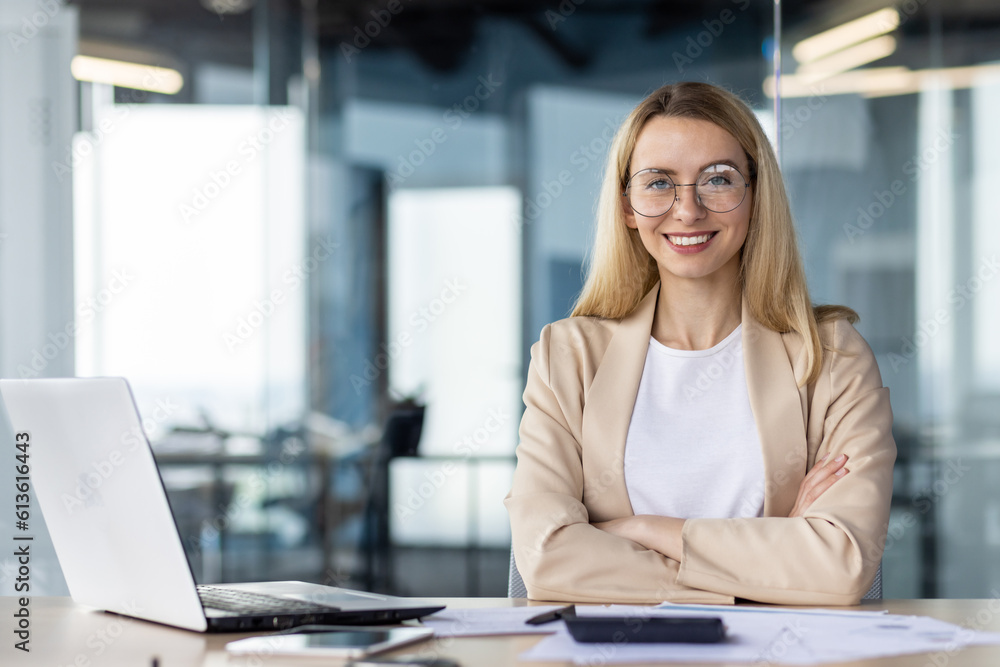 Portrait of a successful and self-confident business woman in a business suit sitting in the office at the desk and looking at the camera smiling with her arms crossed Portrait of a successful and self-confident business woman in a business suit sitting in the office at the desk and looking at the camera smiling with her arms crossed