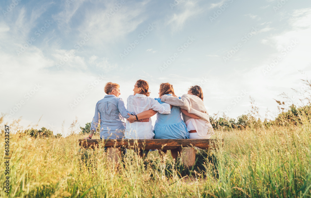 Fototapeta premium Four smiling women dressed light summer clothes harm embracing sitting on the meadow bench during outdoor walking. Woman's friendship, relations, and happiness concept image.