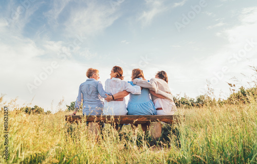 Four smiling women dressed light summer clothes harm embracing sitting on the meadow bench during outdoor walking. Woman's friendship, relations, and happiness concept image.