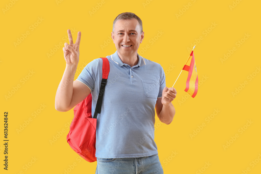 Mature man with flag of Spain and backpack showing victory gesture on yellow background Mature man with flag of Spain and backpack showing victory gesture on yellow background