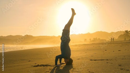 Fotografie SILHOUETTE: Vital young man practising yoga headstand on exotic beach at sunset