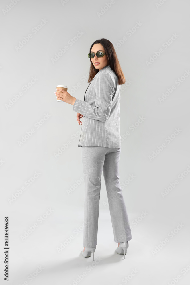Young woman in stylish suit with cup of coffee on light background Young woman in stylish suit with cup of coffee on light background