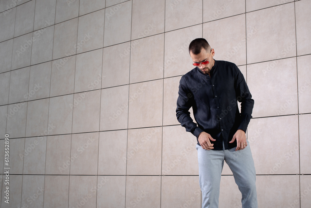 a stylish man in light pants and a black shirt on the background of an empty wall takes off something in sunglasses and looks down a stylish man in light pants and a black shirt on the background of an empty wall takes off something in sunglasses and looks down