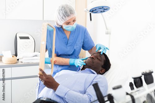 African american male client looking at mirror while qualified beautician examining his face after procedure in medical cosmetology office