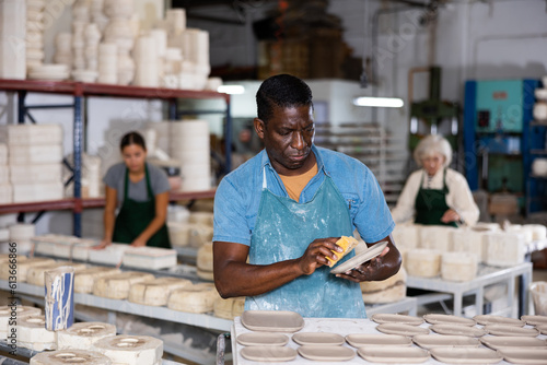 Skillful african-american man ceramist in apron polishing new plates in workshop.